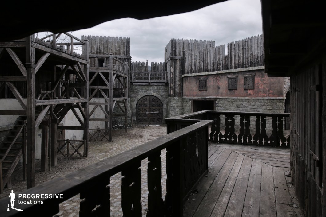 This enclosed fortress courtyard is surrounded by high stone walls topped with dense wooden palisades and interconnected timber walkways. The construction blends raw, functional elements with layered defensive structures, including arched gates, narrow windows, and elevated platforms. The gritty, weathered textures and labyrinthine architecture lend the space an atmosphere of tension and confinement. This location is ideal for filming siege sequences, prison breakouts, covert operations, or military life in historical or fantasy genres.