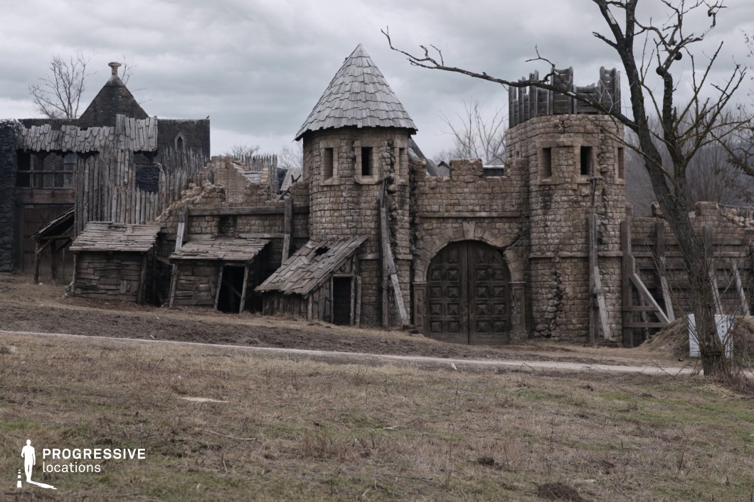 This rugged exterior depicts a fortified medieval outpost with a central arched gate flanked by two cylindrical towers, topped with conical and battlemented structures. The construction utilizes rough-hewn stone blocks and timber supports, conveying a sense of age and structural wear. Adjacent to the gate, deteriorating wooden lean-tos and jagged palisades enhance the rustic, frontier aesthetic. The overall composition is grounded in earthy tones and asymmetrical forms, evoking an atmosphere of historical grit and isolation. This location is particularly suitable for fantasy, war, or post-apocalyptic film productions seeking an authentic, distressed stronghold or rural settlement setting.