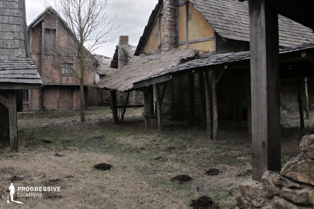 This quiet corner of a rural village features timber-framed buildings with thatched and shingled roofs, worn plaster walls, and a prominent stone chimney. The uneven terrain, scattered patches of grass, and simple wooden structures evoke a sense of everyday medieval life. The mix of textures and building styles creates a convincing, grounded atmosphere. This location is ideal for filming scenes involving rural communities, humble domestic life, or transitional moments in fantasy and historical dramas where characters pass through or seek refuge in modest settlements.