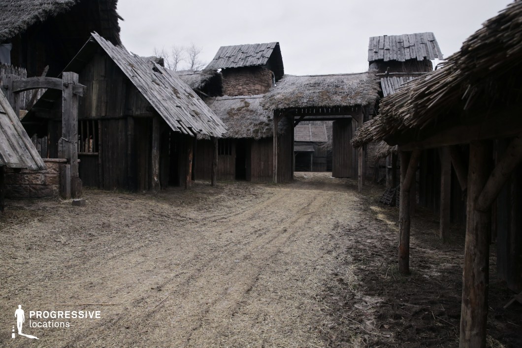 This enclosed hamlet is constructed with rough timber, straw thatching, and clay-reinforced walls, forming a compact, defensive layout centered around a dirt path and a large wooden gate. The hand-hewn textures, irregular rooflines, and rudimentary joinery evoke a harsh, survival-focused environment rooted in an early medieval or tribal setting. The space feels both isolated and self-contained, making it highly effective for scenes in historical dramas, early settlement narratives, or dark fantasy films, especially those depicting frontier life, invasions, or the struggles of pre-feudal communities.
