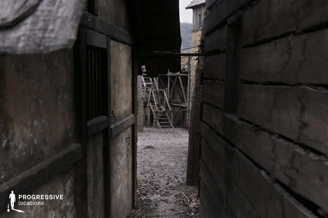 This scene captures a narrow, shadowed passageway between rough wooden and daub buildings, opening onto a rudimentary wooden structure in the background. The textures of aged timber, exposed beams, and packed earth contribute to an atmosphere of gritty realism and modest construction. The confined framing and limited visibility enhance a sense of tension and secrecy. This location is excellent for filming stealthy pursuits, secretive exchanges, or sudden confrontations in genres such as historical drama, low-fantasy, or medieval thrillers.