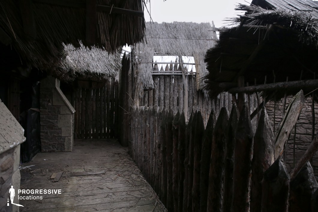This narrow wooden walkway is enclosed by sharpened log palisades and overhung by thatched roofing, creating a compact, defensible corridor within a rudimentary fort or tribal village. The rough textures, exposed wooden framework, and uneven construction evoke a raw, survivalist atmosphere, ideal for portraying early settlements, frontier outposts, or tribal enclosures. This location works especially well for historical or fantasy productions depicting sieges, escapes, hidden movements, or tense confrontations within a fortified stronghold.