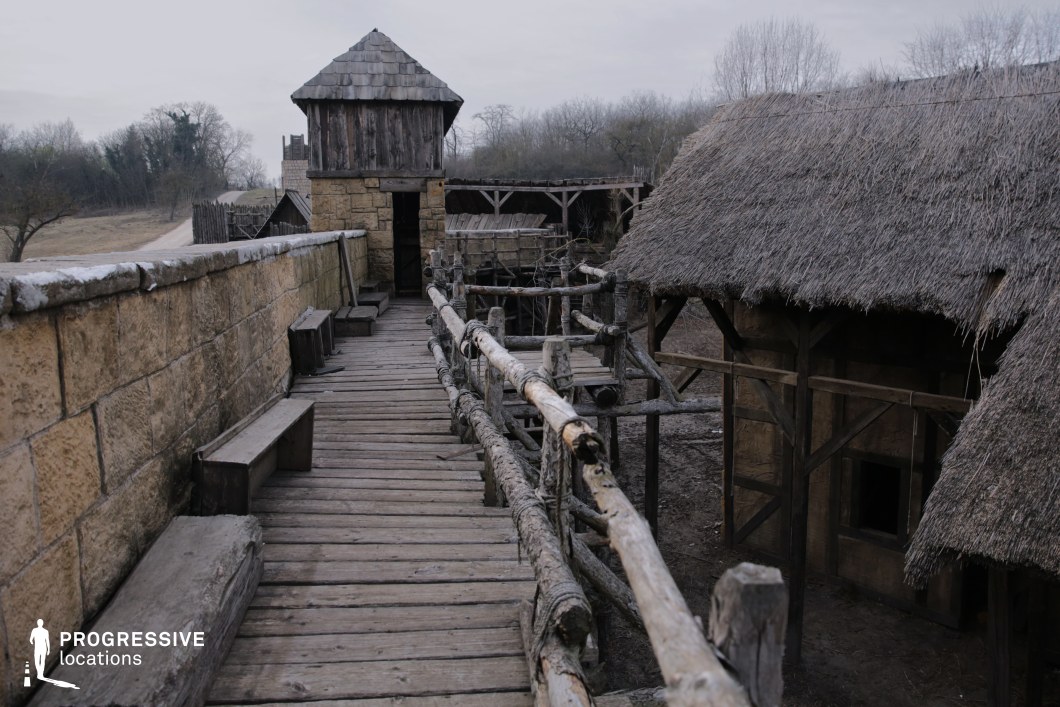 This scene presents a fortified wooden walkway atop a stone wall, leading toward a rustic watchtower clad in timber with a shingled roof. To the right, thatched-roof structures and simple log railings add to the authentic, pre-modern ambiance. The construction is entirely composed of rough natural materials—stone, logs, and reed—conveying a raw, utilitarian style appropriate for a frontier or rural outpost. The elevated path and benches suggest defensive or lookout functionality, and the enclosed configuration offers strong directional perspective. This location is well-suited for historical epics, medieval adventures, or survivalist-themed productions, providing a visually immersive and strategically layered setting.