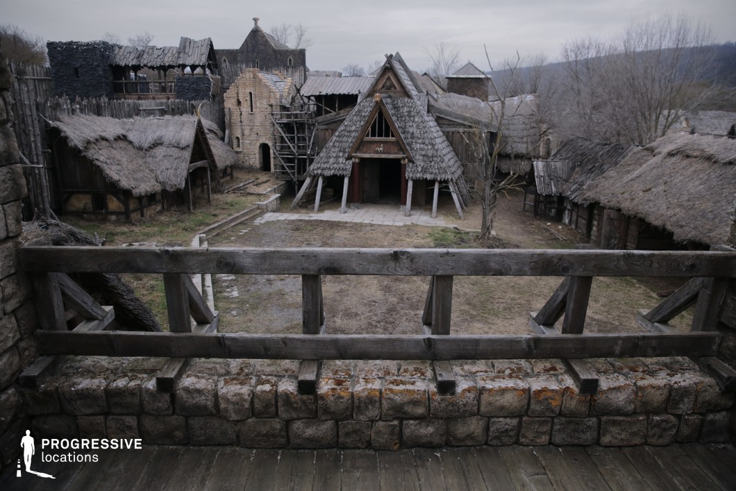 This location depicts a rustic, fortified settlement with steeply pitched wooden roofs, thatched huts, and palisade walls, centered around a triangular timber hall. The elevated viewpoint over a crude stone parapet adds a tactical visual element, enhancing the impression of a defensible tribal or early medieval village. The rough-hewn materials and earthy tones give the space a primitive, immersive atmosphere. This setting is ideal for filming ancient or mythic cultures, tribal councils, or siege scenarios in fantasy epics, historical dramas, or Viking-inspired stories.