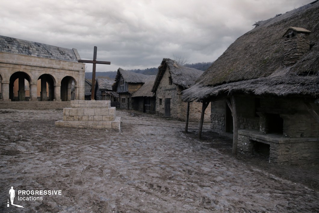 This setting captures a rustic medieval village with thatched stone cottages, a cobbled and muddy central square, and a prominent stone structure with arched openings on one side. A large cross stands on a tiered pedestal at the center, reinforcing the spiritual and communal tone of the space. The raw, earthy textures and architectural simplicity evoke a sense of hardship and authenticity. This location is ideal for historical dramas, folk tales, period epics, or fantasy films, and suits scenes involving peasant life, village gatherings, rituals, or conflict between rural and ecclesiastical powers.