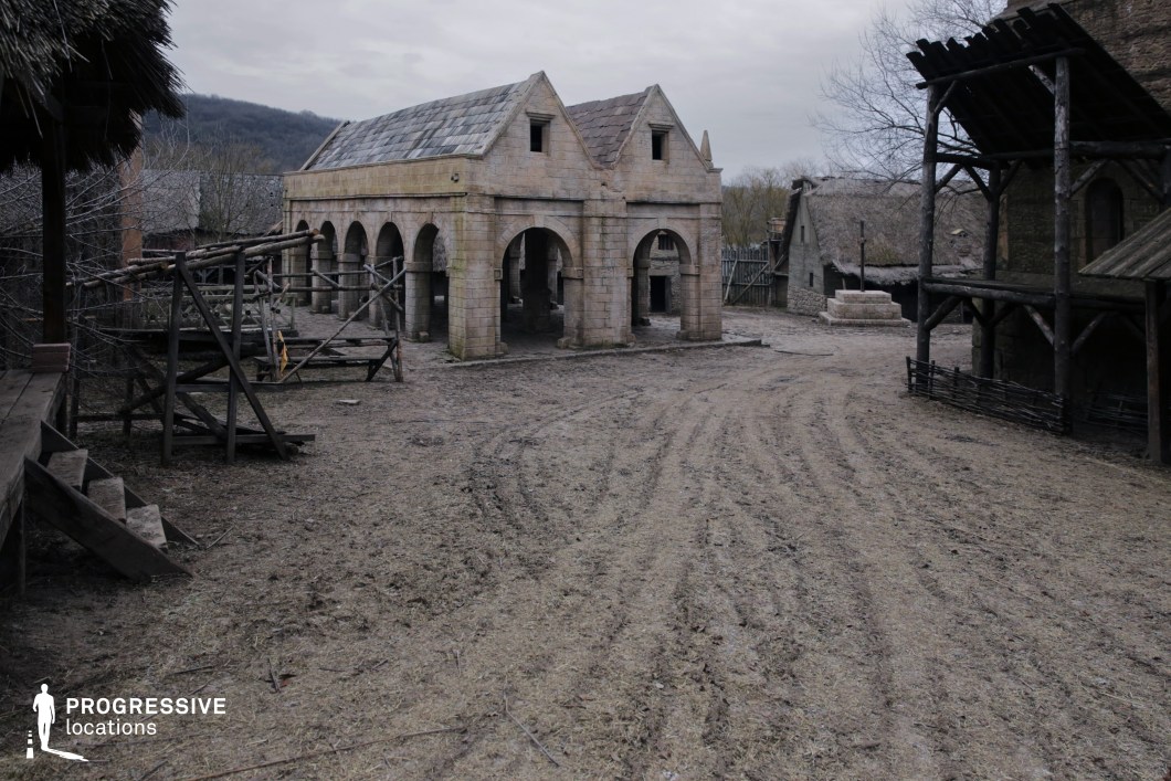This broad, unpaved village square is dominated by a symmetrical stone hall with twin gables and a series of arched openings, evoking a monastic or civic function. Surrounding structures include thatched cottages and wooden scaffolding, enhancing the rural, pre-industrial character of the setting. The natural terrain and weathered architecture contribute to an immersive, gritty atmosphere. This location is ideal for medieval-themed films, fantasy series, or historical reconstructions, perfect for scenes involving market gatherings, trials, or pivotal village confrontations.