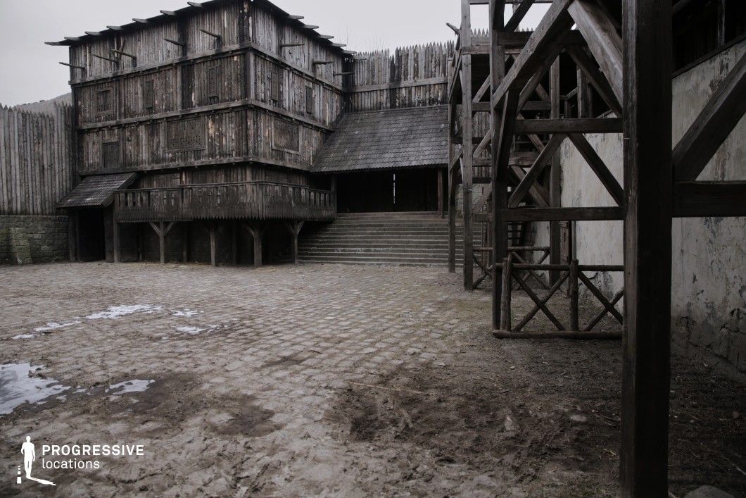 This enclosed courtyard is surrounded by tall palisade walls and imposing timber structures featuring elevated balconies, staircases, and platforms. The ground is uneven with cobblestone and compacted earth, adding a rugged, lived-in texture to the scene. With its fortified appearance and defensible layout, this location is perfect for filming medieval fort scenes, military encampments, or tribal strongholds, especially for action sequences, tense negotiations, or preparations before battle.