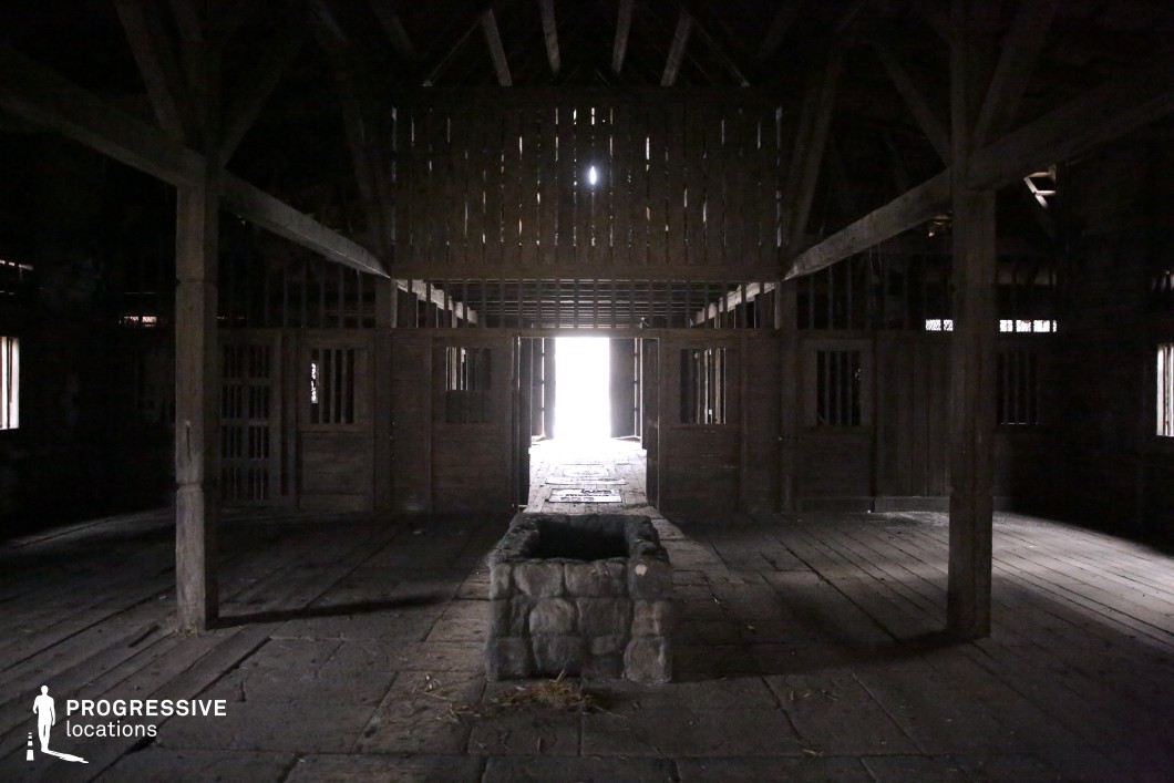 This dimly lit wooden structure features thick support beams, a lofted upper section, and a central stone trough on a rough plank and stone floor. The architecture and layout suggest a traditional stable or barn, with gated compartments and a large doorway casting dramatic light into the otherwise shadowy interior. The space feels aged, utilitarian, and atmospheric. This location is well-suited for historical dramas, rural-themed films, or fantasy stories, ideal for scenes involving secrecy, captivity, nighttime confrontations, or quiet moments of reflection.