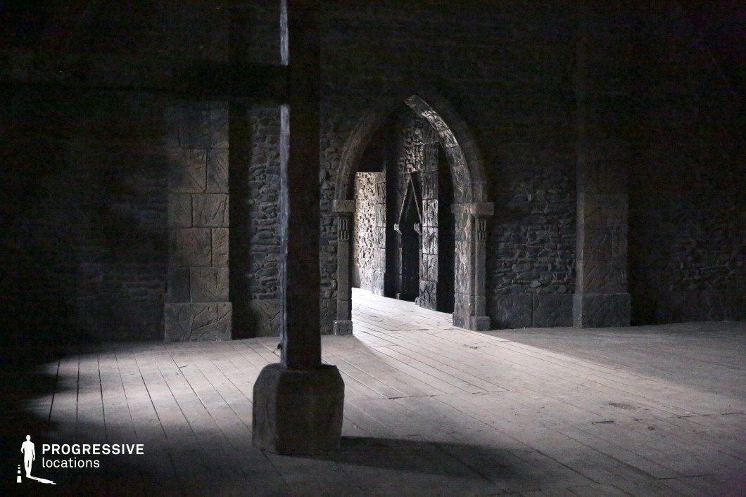 This dimly lit interior features heavy stone walls, pointed archways, and a wide wooden plank floor, creating an austere and ancient atmosphere. The rough-hewn textures and structural simplicity evoke a sense of solemnity and age, while the interplay of shadow and light adds visual drama. The space is adaptable for scenes set in castles, temples, or fortresses, making it ideal for fantasy, medieval, or gothic-themed productions, particularly for rituals, secret meetings, or moments of suspense within sacred or guarded halls.