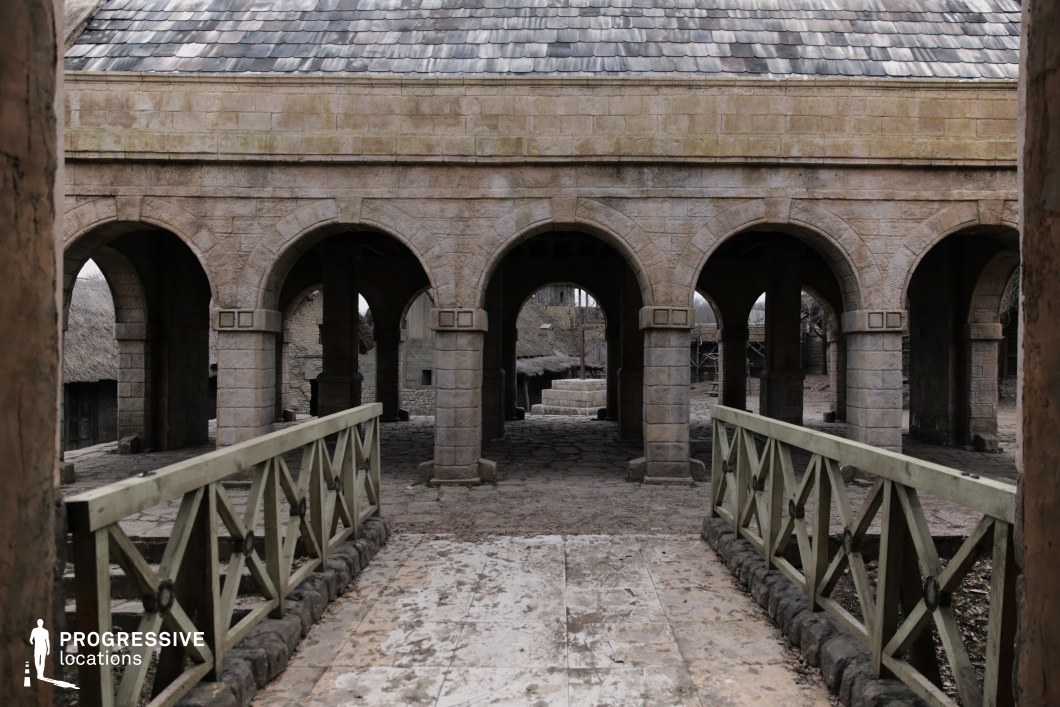 This symmetrical structure features a row of rounded stone arches supported by thick columns, fronted by a short wooden bridge with rustic railings and stone bases. The heavy roof tiles and worn stonework suggest a sacred or administrative function, possibly a chapter house, monastery, or courthouse. The layered arches create a strong sense of depth and order, while the direct axial view through the structure offers visual storytelling opportunities. This location is ideal for historical or fantasy films involving rituals, councils, executions, or clandestine meetings, and lends itself to both tense dialogue scenes and ceremonial sequences.