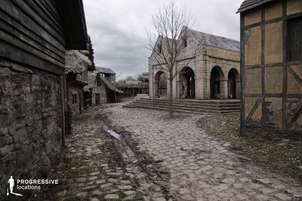 This rustic village setting centers around a worn cobblestone street flanked by timber-framed and stone buildings, leading to a prominent arched stone hall that evokes a temple, market house, or village courthouse. The textures of rough stone, wood, and thatch create a convincingly aged and immersive environment. The open layout and tiered steps in front of the hall provide natural staging areas for dramatic scenes. This location is ideal for medieval or fantasy films, suitable for town gatherings, trials, market scenes, or moments of public tension and revelation.