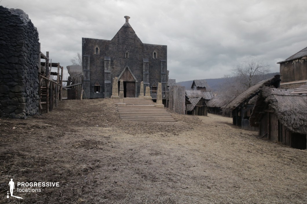 This scene presents a rugged village dominated by a striking central stone temple or hall, accessed by a broad staircase and flanked by wooden and thatched structures. The earthy terrain and raw construction materials evoke a sense of early civilization or tribal society. The architectural contrast between the imposing, monolithic temple and the surrounding humble dwellings creates visual hierarchy and narrative potential. This location is highly suited for filming fantasy epics, dark historical sagas, or post-apocalyptic tales, particularly scenes involving rituals, village life, or power struggles rooted in ancient or mythical cultures.