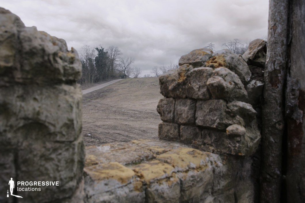 Seen through a narrow gap in a weathered stone wall, this view reveals an open, sloped landscape with a dirt path leading into sparse woodland. The rough masonry and partially broken opening suggest a defensive or concealed vantage point, ideal for surveillance or ambush scenes. The setting conveys isolation and vulnerability, making it perfect for tense moments in war films, fantasy adventures, or medieval dramas—especially scenes involving scouts, sentries, or sudden threats emerging from the terrain beyond.