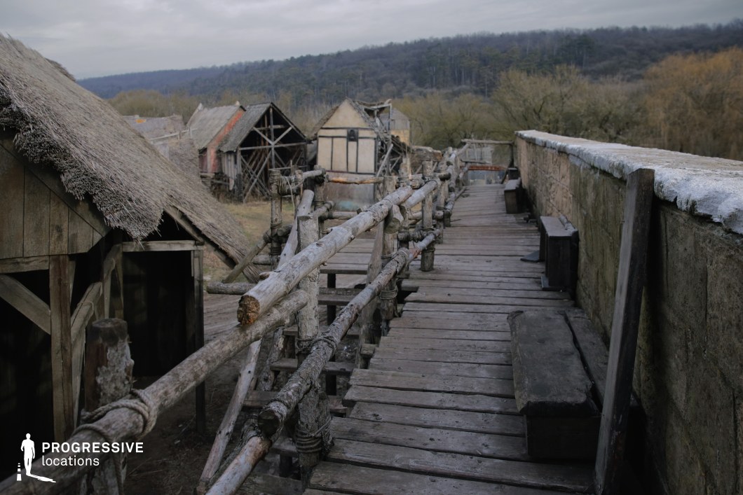 This elevated wooden walkway runs along a stone wall and overlooks a cluster of primitive, timber-framed and thatched buildings set against a backdrop of dense forest. The rough-hewn railings, tied with rope, and the weathered construction evoke a frontier or early medieval settlement. This location is ideal for filming historical epics, fantasy adventures, or survival-themed stories, particularly for scenes involving guards on patrol, village life in remote regions, or dramatic standoffs above rural settlements.