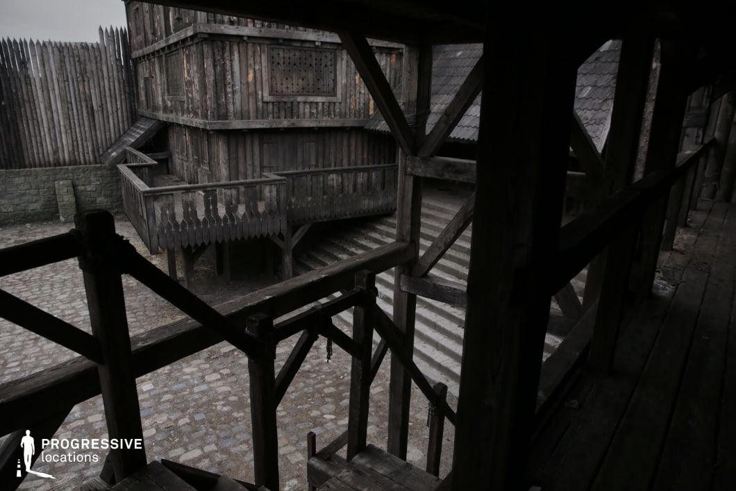 This scene features a fully enclosed wooden compound with defensive palisade walls, a timber-framed structure with overhanging balconies, and multi-level staircases surrounding a cobblestone courtyard. The dark wood construction and narrow passageways convey a sense of confinement and tactical design, typical of strongholds or garrisons. The layered elevation and tight geometry make this an ideal setting for suspenseful encounters, stealth sequences, or power plays in historical, fantasy, or post-apocalyptic genres—especially scenes involving ambushes, judgment platforms, or internal conflict within a fortified enclave.