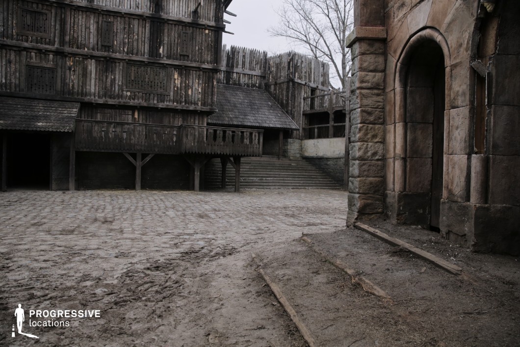 This gritty medieval courtyard is framed by dark timber buildings elevated on stilts and supported by rough stone and wood reinforcements. The courtyard floor transitions from cobblestone to packed earth, with visible wear enhancing its lived-in character. The architectural palette includes vertical wooden planks, sloped shingled roofs, and fortified palisade elements, contributing to a fortified village or garrison ambiance. On the right, a heavy stone doorway adds architectural contrast and a sense of permanence. The layered elevation and varied materials create a highly atmospheric setting, ideal for fantasy, historical, or post-apocalyptic productions requiring a fortified village, barracks, or transitional urban zone.