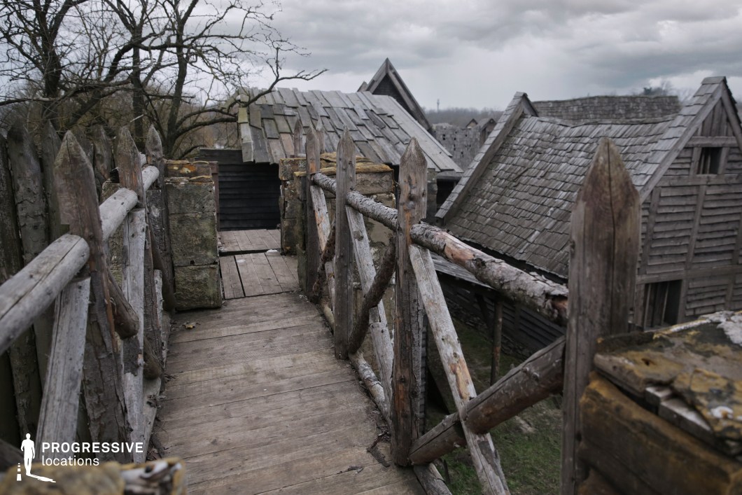This narrow wooden walkway, framed by rough-hewn timber railings and spiked palisade posts, offers an elevated view across a village of steep, shingled rooftops. The surrounding structures, built from aged planks, evoke a sense of isolation and age. This location is well-suited for historical or fantasy productions, ideal for scenes involving lookouts, stealthy movement across fortifications, or tense moments of observation before conflict.