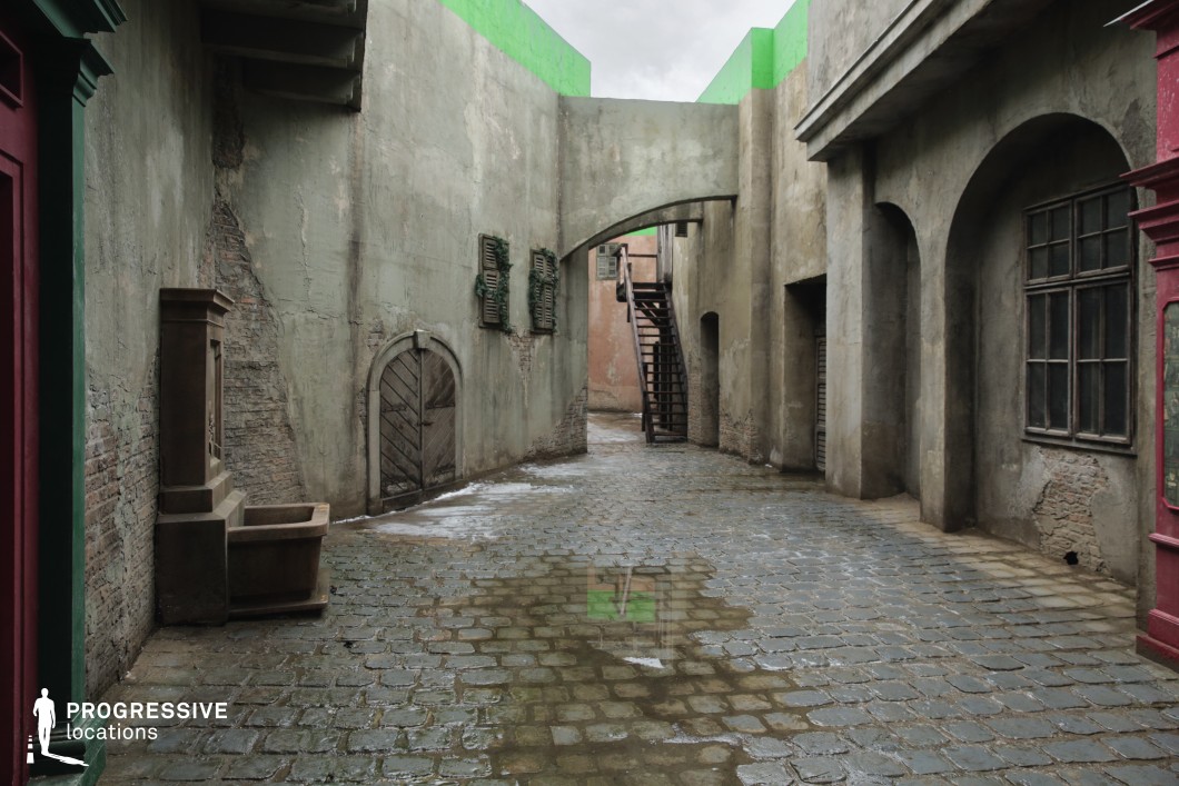 This narrow alleyway features aged, weathered facades with a mix of rough concrete and exposed brick textures, creating a distressed and raw visual atmosphere. The cobblestone pavement adds irregularity and historical texture to the ground surface, complemented by water stains that enhance the worn look. Architectural elements include arched windows and passageways, wooden shutters in disrepair, a modest corner fountain structure, and an external wooden staircase leading to an upper level, which adds vertical layering to the space. The high walls and overpass arch create a semi-enclosed feeling, intensifying the sense of confinement. This location is highly versatile for filming, particularly suited for historical dramas, period thrillers, or dystopian urban scenes, offering a visually immersive and gritty setting.