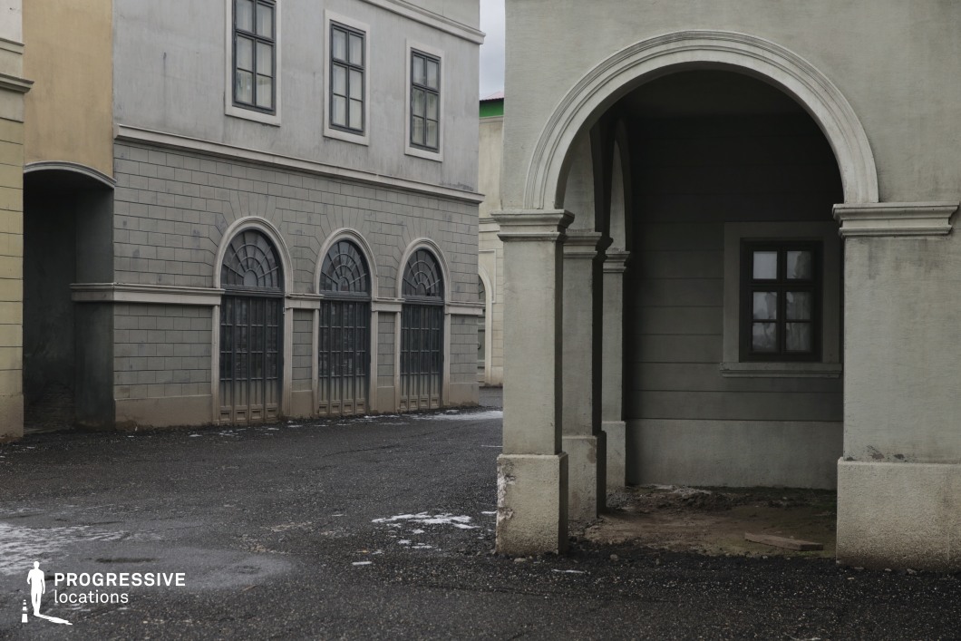 This location features muted grey-toned façades with symmetrical neoclassical elements such as arched windows, deep-set frames, and clean columned archways. The sharp lines and uniform surfaces evoke a stylized, somewhat austere urban environment. The architectural design lacks ornamentation, creating a mood of quiet tension or institutional rigidity. This location is well-suited for filming noir thrillers, political dramas, or dystopian stories. Its structured geometry and shadow-filled passageways provide ideal backdrops for suspenseful chases, introspective character moments, or bureaucratic power plays.