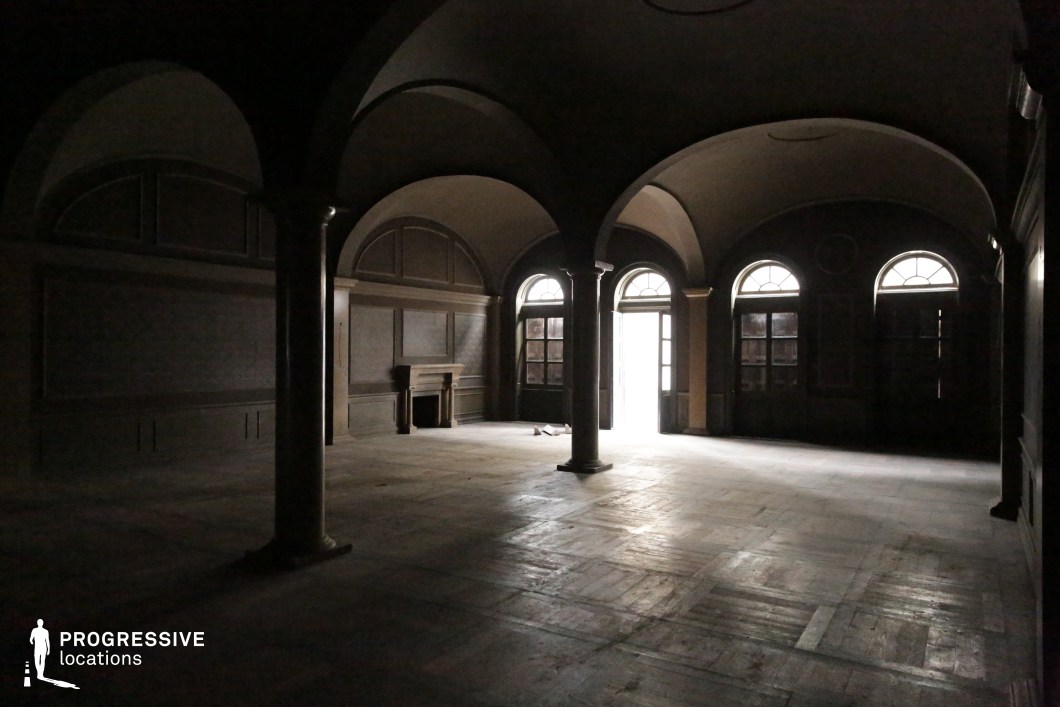 This interior space features a grand arched ceiling supported by classic cylindrical columns, evoking an atmosphere of faded opulence. The room is characterized by subdued, dark wood tones throughout the flooring, wainscoting, and structural elements, complemented by large arched windows and doors that allow limited natural light to penetrate. A non-functional fireplace is centrally positioned along one wall, enhancing the historical character of the space. The parquet floor, though worn, contributes to the room's textured and atmospheric quality. The overall structure creates a rhythm of arches and shadow that lends itself to moody and dramatic visuals. This location is particularly well-suited for filming period pieces, thrillers, or introspective dramatic scenes where architectural depth and ambient shadow are crucial to visual storytelling.