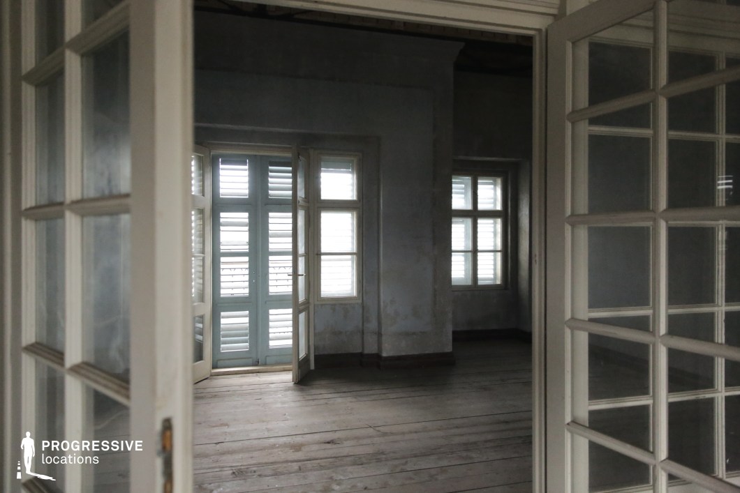 This location presents an empty, multi-room interior space defined by aged wooden flooring, muted grey walls, and classical white-framed glass doors. The visible structure includes a set of shuttered double windows and doors that allow diffused natural light to softly filter through, contributing to a subdued, introspective atmosphere. The wooden floorboards exhibit a worn texture that enhances the historic and unpolished aesthetic of the space. The architecture favors traditional proportions with moderate ceiling height and thick interior walls that segment the space into distinct but visually connected rooms. The camera position—angled from an ajar doorway—emphasizes depth and layered perspectives, ideal for cinematic compositions. This location is highly suitable for film scenes requiring a nostalgic, melancholic, or suspenseful tone, making it fitting for dramas, psychological thrillers, or period pieces that benefit from intimate, atmospheric interiors.
