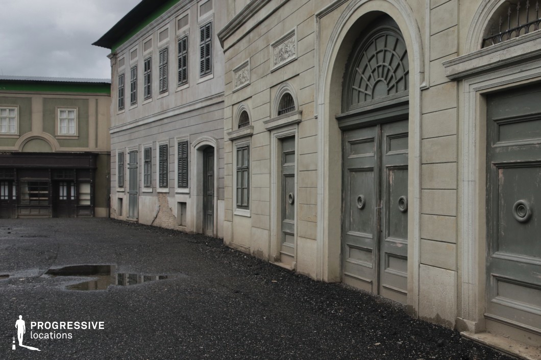 This location showcases a courtyard enclosed by multi-story classical-style buildings featuring symmetrical facades, tall shuttered windows, and arched doorways. The walls are finished in muted tones of grey and green, with visible patina and peeling plaster that contribute to an aged, authentically worn aesthetic. Architectural details include rectangular and arched stone frames, modest reliefs, and traditional cornices that evoke 19th-century European urban architecture. The textured asphalt courtyard, complete with uneven surfaces and water puddles, reinforces the slightly neglected ambiance. The composition emphasizes geometric repetition and perspective, with the camera positioned at an angle that draws attention to the linearity and architectural rhythm of the façades. This site is ideal for period films, historical dramas, or productions requiring a neutral European street setting, offering versatility for both urban scenes and introspective, mood-heavy narratives.