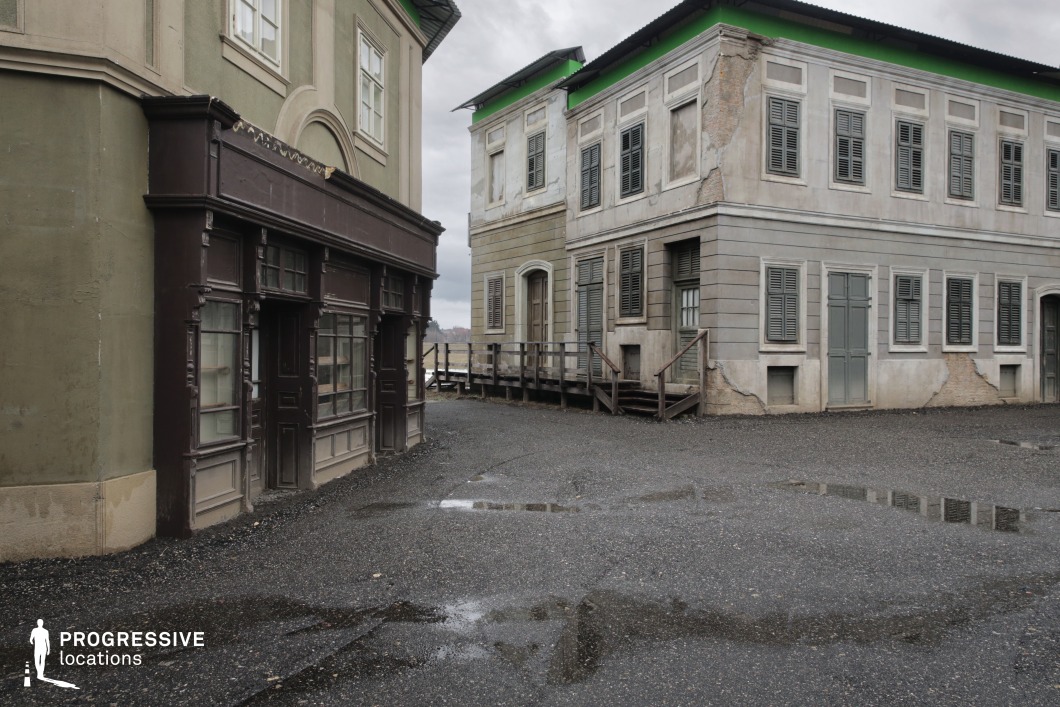 This enclosed corner of a backlot set features Hungarian-style urban architecture with two- to three-story plastered buildings, showcasing a deliberately aged and distressed finish. The façades exhibit muted tones of olive green, beige, and grey, with flaking paint and exposed brick details enhancing the worn aesthetic. The left structure includes a dark brown storefront with carved wooden framing and large display windows, suggestive of early 20th-century commercial design. Shuttered windows dominate the right-side building, and wooden stairs lead up to mismatched doorways, adding textural variety and dimensional layering. The gravel ground and visible puddles reinforce the rugged, utilitarian atmosphere. This location is ideal for period films, crime dramas, or dystopian stories, offering a flexible and immersive environment for scenes that require authenticity, decay, or old-world urbanity.