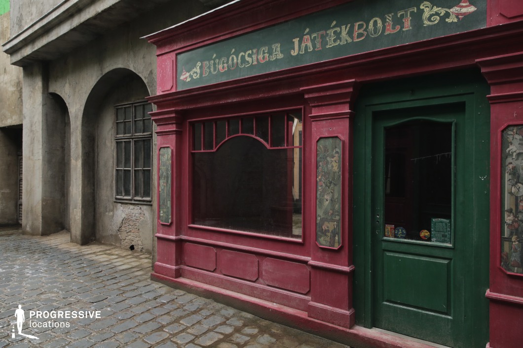 This location presents a vividly colored, old-fashioned storefront framed by worn, grey plaster buildings, creating a compelling contrast between decorative charm and raw urban texture. The toy store façade features a dark green door and display window bordered by deep red wooden paneling, detailed with painted illustrations in narrow vertical frames. The aged signboard and slightly distressed paintwork enhance the nostalgic aesthetic. Surrounding structures exhibit exposed stonework, arched supports, and weathered window frames, reinforcing the timeworn atmosphere. The cobblestone street adds to the period authenticity and offers rich texture underfoot. This space is particularly suitable for filming scenes set in historical or fantasy contexts, evoking early 20th-century or Central European urban environments, and works well for genres such as drama, mystery, or period fantasy.