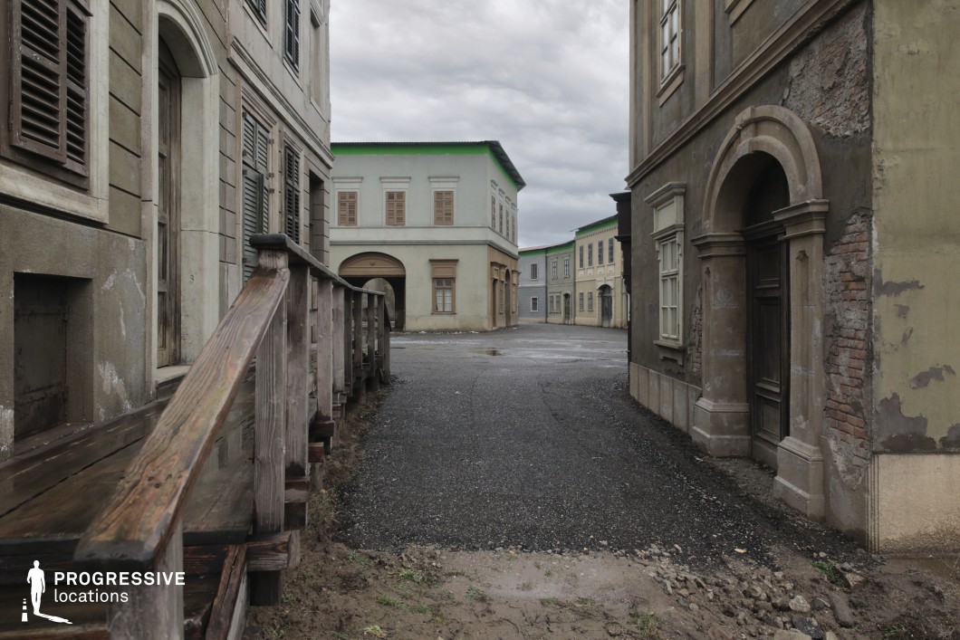 This image presents a cinematic street composed of distressed façades, worn plaster, exposed brickwork, and faded architectural detailing, evoking a bygone European cityscape. The uniform yet deliberately aged appearance of the buildings creates a cohesive and immersive historical environment. The spatial layout with narrow alleys, wide-open intersections, and varied textures enhances the visual storytelling potential. This location is excellent for filming war dramas, post-apocalyptic narratives, or historical fiction, offering versatile settings for chase scenes, quiet moments of reflection, or sequences portraying social decay and transformation.