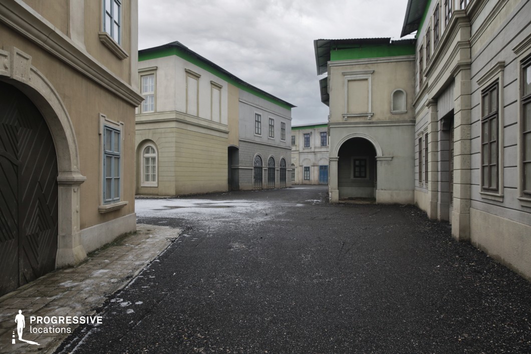This location features a spacious, enclosed urban set composed of two-story buildings in muted beige and gray tones with consistent neoclassical design motifs. The facades are characterized by arched doorways, pilasters, symmetrical window arrangements, and simplified cornices, contributing to a stylized yet generic European cityscape. The structures lack intricate detailing, creating a versatile and adaptable environment. A distinctive feature is the uniform green trim along the building tops, which may suggest its use as a film set with post-production flexibility in mind. The ground is paved with dark asphalt and stone, contrasting with the soft-toned buildings and enhancing depth. The angular perspective and clean sightlines make this location suitable for tracking shots and wide establishing frames. This space is particularly well-suited for controlled film productions, including historical dramas, thrillers, or fictional urban settings, offering a blank canvas for varied creative interpretations.