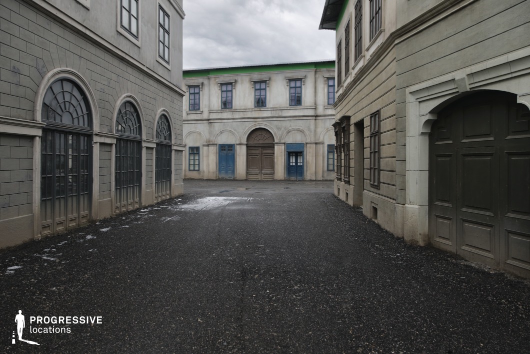 This backlot location represents a stylized European alley or narrow street framed by classical façades with muted stone textures and subtle neoclassical detailing. The façades display repeated architectural motifs like rectangular and arched windows, flat pilasters, and recessed panels. The surfaces are treated in a weathered, grey-beige tone that contributes to an aged, lived-in appearance. The spatial arrangement forms a funnel-like corridor leading to a two-story building with a tripartite doorway—arched in the center, flanked by rectangular frames—establishing a strong visual endpoint. Green chroma key edging on the rooflines confirms the modular and customizable nature of the set. This space is ideal for controlled film shoots requiring intimate cityscapes or back-alley perspectives in historical, mystery, or fantasy genres, where set extension and stylized production design are key.