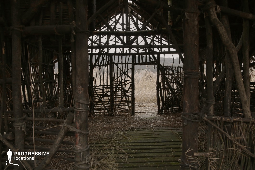 This interior view reveals a hand-crafted wooden shelter made from interwoven branches and rough-hewn timber, secured with visible cord binding. The walls are open and irregular, allowing filtered views of the surrounding tall grassland and distant hills through the loosely constructed grid. The structure’s uneven texture and natural materials create a raw, archaic aesthetic, reinforced by the moss-covered wooden floorboards and scattered straw underfoot. The muted, earthy tones contribute to a sense of simplicity and ancient functionality. This location is ideal for primitive or prehistoric-themed productions, offering an immersive backdrop for historical, survival, or fantasy scenes that require authenticity and elemental visual language.