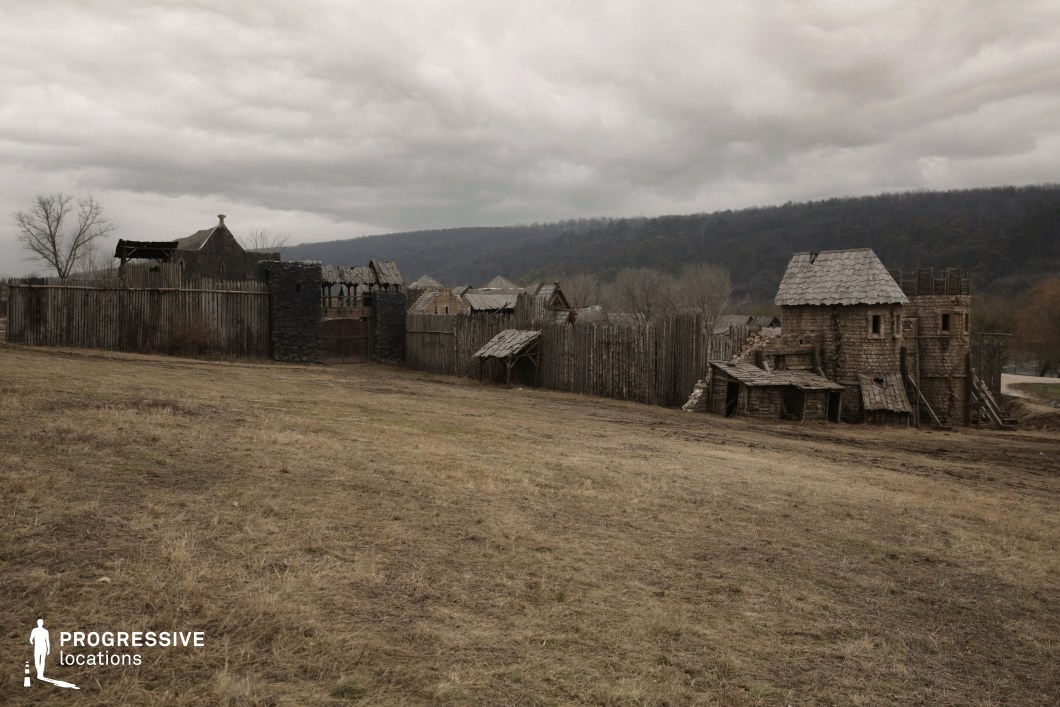 This location features a rustic, enclosed settlement designed in medieval architectural style, situated on a slightly sloped, open terrain with a wooded hillside in the background. The perimeter is defined by a tall, uneven wooden palisade interrupted by a fortified stone gate and watchtower, adding to the sense of historical realism. Inside the fence, timber and stone structures with steep, shingled roofs exhibit intentionally weathered surfaces and irregular construction, enhancing the handcrafted, archaic aesthetic. The materials—primarily raw wood and coarse stone—emphasize a pre-modern, utilitarian atmosphere. The setting’s openness and surrounding natural elevation provide excellent depth and compositional layering for wide, panoramic shots. This location is ideal for historical or fantasy productions, offering an immersive backdrop for action scenes, period storytelling, or epic dramas requiring an authentic, village-scale medieval environment.