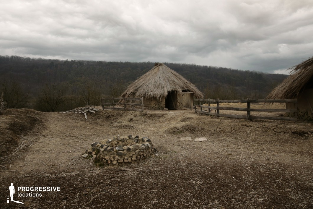 This remote rural location features traditional round huts with thick, conical reed-thatched roofs and walls constructed from natural clay and timber, evoking a prehistoric or early agrarian settlement. The terrain is dry and uneven, with patches of straw and dirt dominating the ground cover, and a primitive circular stone structure in the foreground suggests a fire pit or communal feature. The wooden fencing is irregular and weathered, contributing to the handmade, timeworn aesthetic. In the background, densely wooded hills form a continuous tree line that encloses the scene and enhances the isolation of the setting. This site is well-suited for historical or ethnographic productions, primitive-era reenactments, or documentary-style shoots due to its authentic construction materials, untouched natural surroundings, and strong visual separation from modern elements.