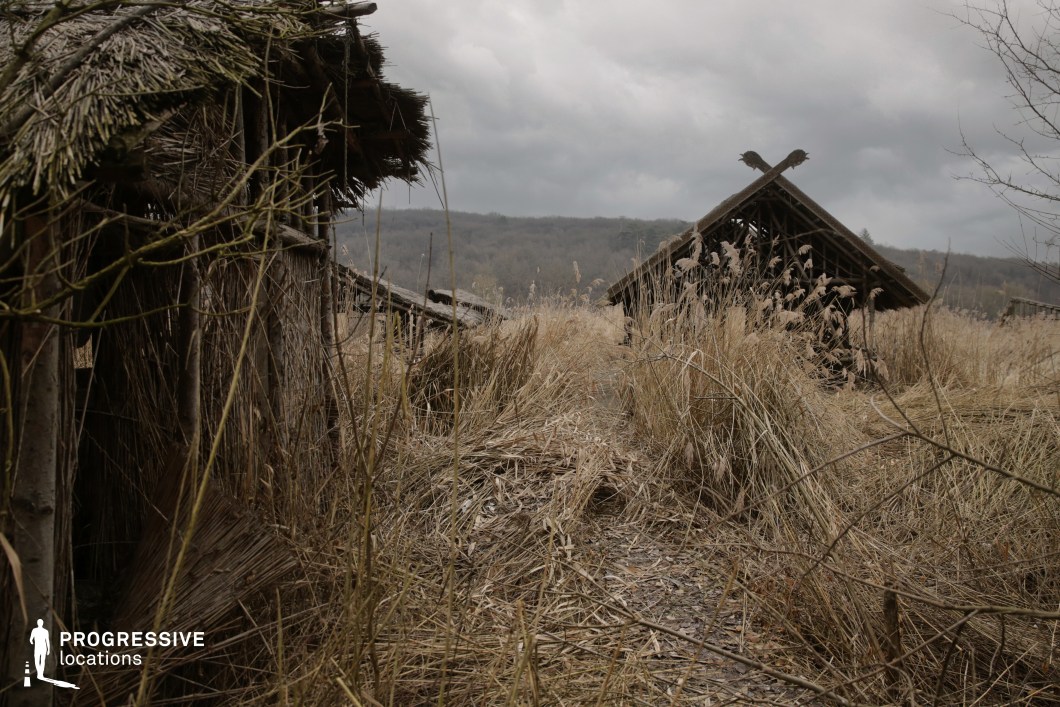 This location features deteriorating reed huts scattered among dense, unkempt grasses and framed by a backdrop of wooded hills. The scene is dominated by earthy textures and tangled organic forms, giving the impression of abandonment and slow reclamation by nature. The rough construction of the structures, coupled with their weathered appearance, evokes a timeless and remote atmosphere. This setting is ideal for prehistoric tales, post-apocalyptic narratives, or folk horror. It offers powerful visual potential for scenes of solitude, survival, or eerie mystery unfolding in a forgotten or untouched world.