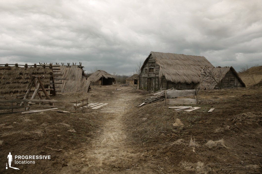 This rustic location features a series of wooden and thatched-roof structures arranged along a dirt path, evoking a pre-modern rural settlement or reconstructed historical village. The buildings are constructed with rough-hewn timber, weathered planks, and straw roofing, emphasizing hand-crafted techniques and functional simplicity. The terrain is uneven and primarily composed of dry, packed earth with sparse vegetation, reinforcing the raw, unrefined atmosphere. The layout is open yet defined by natural barriers and clustered architectural elements. The visual composition benefits from the linear arrangement of huts, which draws the viewer’s eye into the depth of the setting. This site is particularly well-suited for historical reconstructions, early civilization depictions, or productions seeking an archaic, agrarian aesthetic.