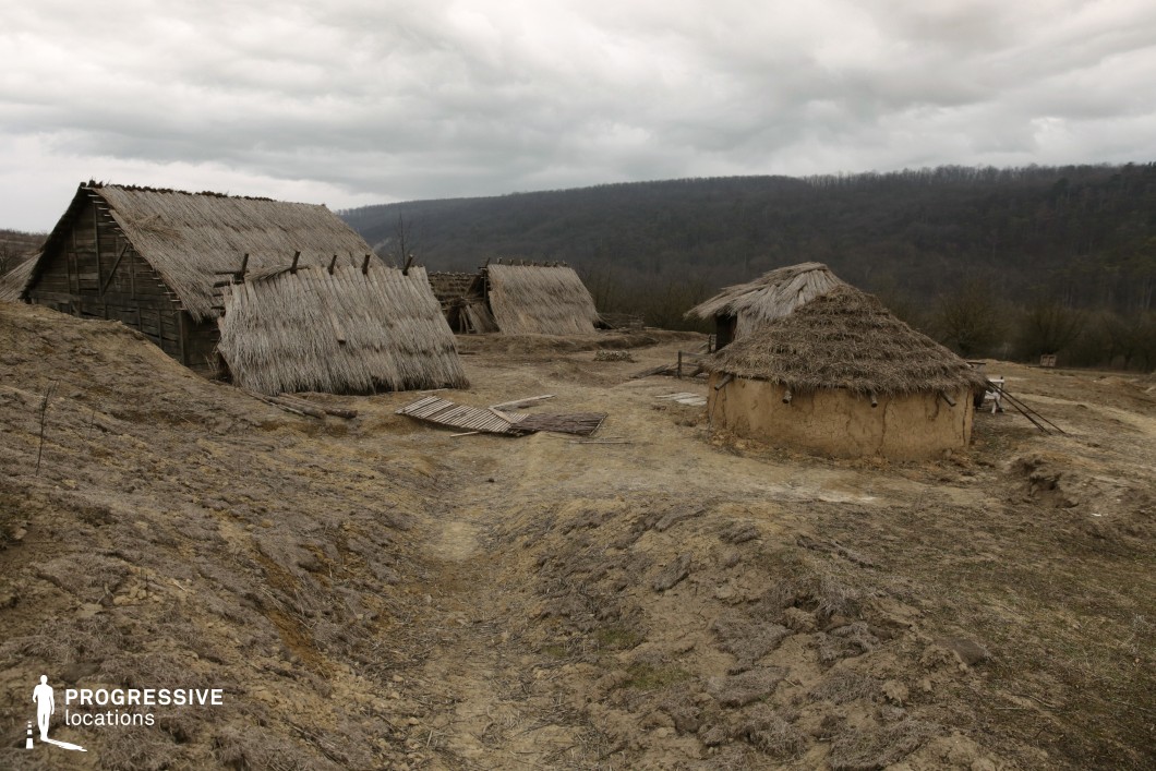 This remote location features a cluster of rudimentary huts constructed from natural materials such as thatch, mud, and timber, set against a sloping, uneven hillside. The structures vary in form, with both conical and gabled roofs, all showing signs of wear, reinforcing the archaic and utilitarian aesthetic. The ground is largely barren, consisting of compacted earth and patches of dry vegetation, and the layout of the village is informal and organically arranged. Surrounding the site is a backdrop of forested hills, providing a sense of isolation and natural enclosure. The authenticity and rawness of the environment make it particularly suitable for historical reconstructions, prehistoric or early tribal settings, or survivalist narratives, offering an immersive backdrop for productions requiring a primitive and untouched atmosphere.