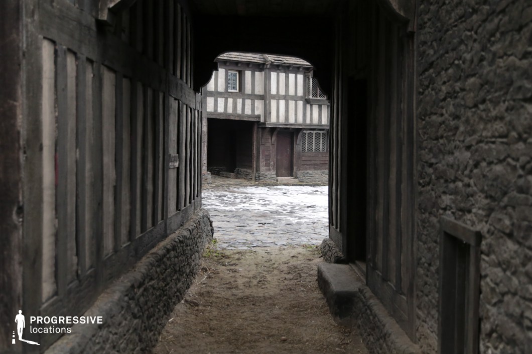 This scene captures a narrow, shadowed passageway framed by rough stone and weathered timber walls, opening into a cobbled courtyard surrounded by traditional half-timbered buildings. The contrast between the dark alley and the brighter square beyond adds natural depth and drama. This authentic, period-specific setting is ideal for filming historical dramas, medieval fantasies, or period thrillers, particularly for scenes involving secret meetings, pursuits through town, or daily life in an old European village.