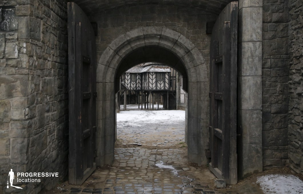 A robust arched stone gateway opens into a narrow cobbled path leading toward half-timbered Tudor-style buildings, creating a layered historical setting. The juxtaposition of fortified stonework and medieval urban architecture suggests a transition from a defensive stronghold to a town or village center. The heavy wooden doors and uneven paving add authenticity and texture. This location is ideal for medieval or fantasy scenes involving town entrances, guarded passages, or transitions between castle and village, making it perfect for dramas, historical action films, or fantasy quests.
