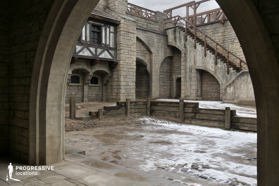 This location features a stone-walled courtyard surrounded by fortified structures, arched doorways, wooden fencing, and a prominent staircase leading to an upper rampart. The blend of stone masonry and timber framing evokes a medieval fortress atmosphere, enhanced by the structured symmetry and layered vantage points. It provides an excellent backdrop for historical epics, fantasy series, or adventure films, ideal for castle life scenes, confrontations, royal processions, or strategic military gatherings.