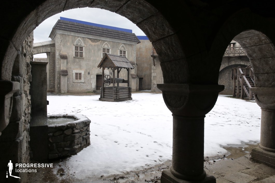 This location presents a stone-built medieval courtyard, framed by arched colonnades and rustic architecture, with weathered plaster façades and wooden elements that suggest historical authenticity. The central well structure enhances the period atmosphere, while the layered stonework and exposed wooden staircases contribute to the sense of age and realism. The scene is ideal for filming historical dramas, fantasy series, or period adventures, particularly for scenes set in a fortified town, castle courtyard, or monastic compound.