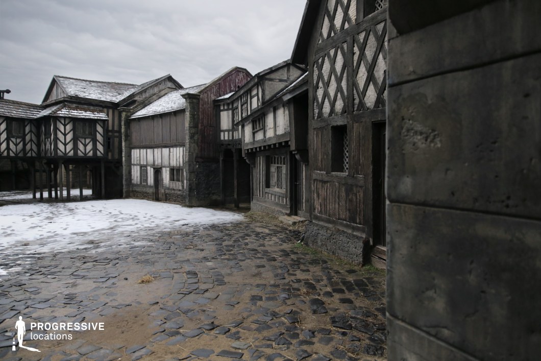 This location presents a convincingly aged village square bordered by timber-framed buildings with dark wooden beams and whitewashed infill, characteristic of late medieval to early modern English architecture. The uneven stone pavement and slightly irregular building facades enhance the authenticity, while the narrow layout suggests a compact and practical historical settlement. Its immersive period look makes it highly suitable for filming historical dramas, fantasy tales, or folklore-inspired stories, especially those requiring village life, market scenes, or confrontations in a rustic public space.