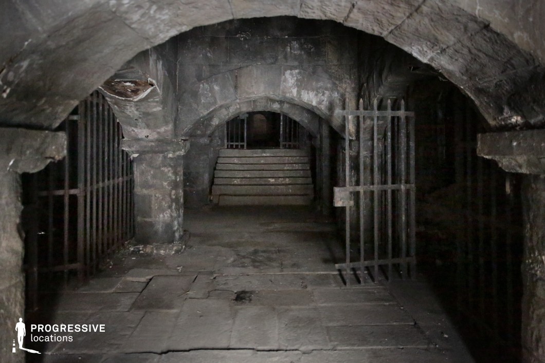 This location features thick, timeworn stone walls, vaulted ceilings, and heavy iron bars embedded in arched alcoves, creating a sense of confinement and foreboding. The central corridor, framed by symmetrical gates and leading toward a shadowy stairway, adds depth and a mysterious focal point. Its dim, dungeon-like atmosphere makes it ideal for filming historical dramas, medieval fantasy, horror, or thriller genres, especially for scenes set in prisons, torture chambers, hidden crypts, or secret lairs.