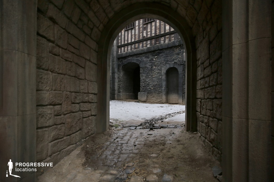 This image captures a narrow arched passageway constructed from regular-cut stone blocks, opening into a walled courtyard with darker masonry and wooden architectural detailing. The transition from the inner corridor to the outer space is emphasized by the contrasting stone textures and shadow gradients, adding visual depth and framing. The cobbled ground continues through the arch and subtly blends with the surrounding packed earth and masonry. The rear structure features a combination of exposed timber framework and irregular dark stone, contributing to a moody, medieval aesthetic. The enclosed geometry and layered materiality make this setting highly suitable for fantasy, historical, or mystery genres, particularly for transitions, reveals, or contained confrontational scenes.