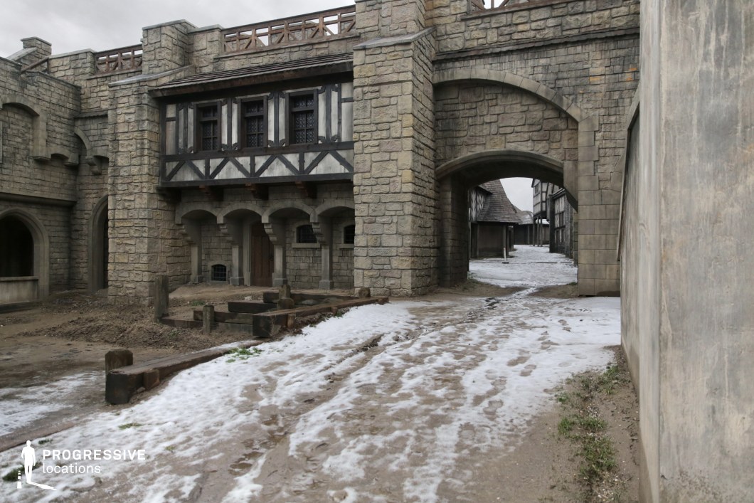 This scene presents a robust stone gatehouse flanked by timber-and-stone buildings, combining fortified architecture with Tudor-style upper levels. The arched entryway leads into a winding street lined with aged façades, suggesting a well-defended urban perimeter or castle courtyard. The large stone blocks, defensive design, and elevated walkway give the area a sense of authority and history. This location is ideal for fantasy series, medieval adventures, or historical dramas featuring town entrances, castle exteriors, or moments of confrontation, patrols, or siege preparations.