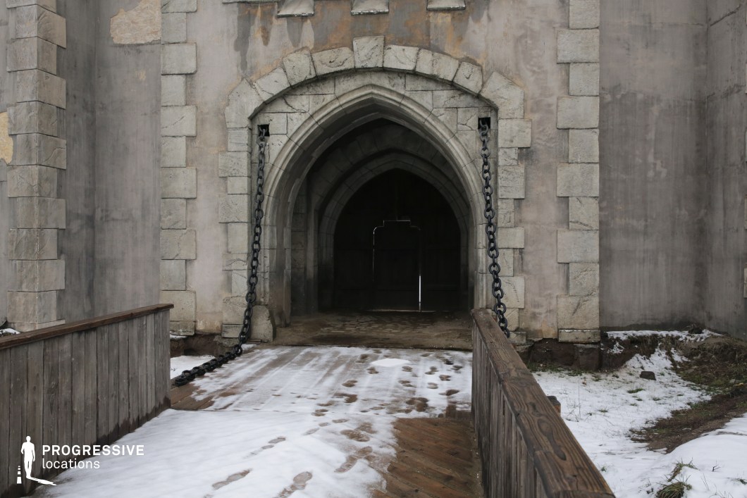 This imposing stone entrance features a classic pointed archway framed by heavy masonry blocks and flanked by large iron chains that suggest a functional drawbridge. The wooden walkway adds texture and historical realism, while the receding dark tunnel behind the gate enhances the sense of depth and mystery. The worn surfaces and stark geometry convey strength and age, making this location ideal for period or fantasy productions. It’s perfectly suited for scenes involving castle entrances, sieges, royal arrivals, or moments of confrontation at a fortified stronghold.