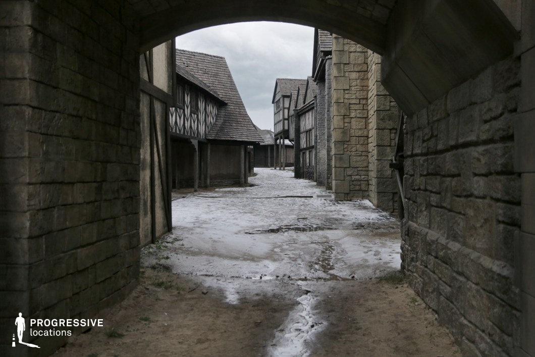 This scene frames a cobbled medieval-style street through a heavy stone archway, creating a strong sense of enclosure and transition. The surrounding buildings feature a mix of rough-cut stone and exposed timber framing, consistent with a fortified town or village from the Middle Ages. The perspective draws the eye down the street, enhancing depth and visual storytelling. This location is particularly well-suited for historical or fantasy genres, perfect for scenes involving city entrances, guarded passages, ambushes, or dramatic character reveals.