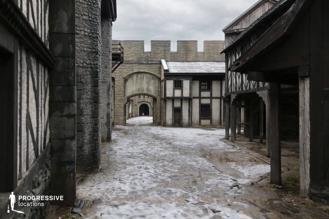 This narrow cobbled street is framed by timber-framed and stone-clad structures, leading the eye through a sequence of arched gateways and culminating in a crenellated defensive wall. The layered architectural elements and slight asymmetries give the space an authentic, timeworn feel, suggestive of a fortified town or city quarter. The sense of enclosure and depth makes this location ideal for filming chase sequences, secret meetings, or ambushes in historical, fantasy, or post-apocalyptic settings, particularly where a sense of confinement and tension is needed.