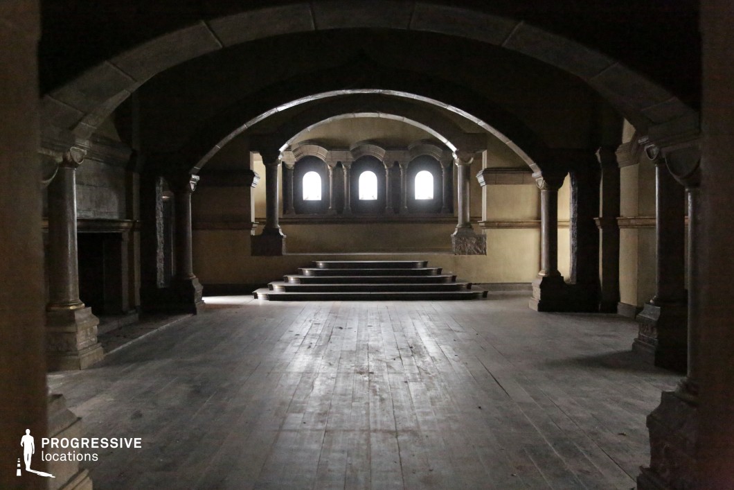 This grand interior space features a rhythmic series of heavy stone arches and columns, leading the eye toward a raised apse framed by ornate Romanesque-style windows. The symmetry, geometric layout, and worn wooden flooring create a solemn and timeless atmosphere. The layered steps and central perspective add a sense of progression and focus, making this location ideal for dramatic entrances, rituals, or confrontational dialogue. It is especially suitable for historical dramas, fantasy epics, secret society scenes, or gothic thrillers requiring a mysterious and stately setting.