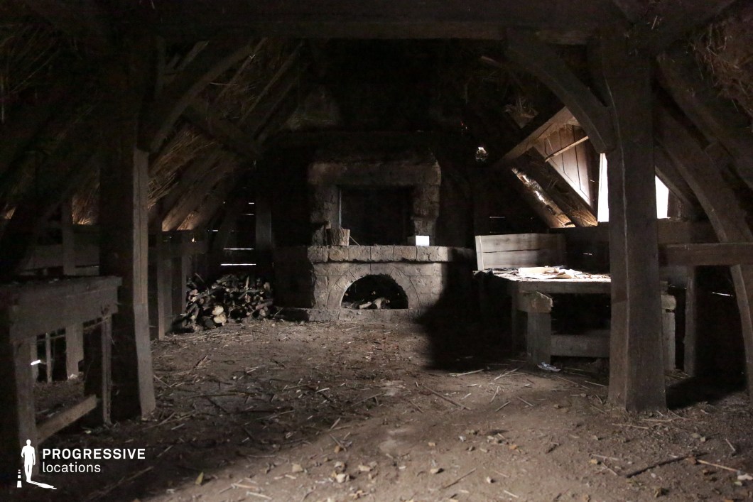 This dimly lit space showcases a rough-hewn wooden structure with a steeply pitched thatched roof and a central stone fireplace built into the back wall. The earth floor, exposed beams, and scattered firewood emphasize the raw, utilitarian nature of the setting. Its primitive and authentic atmosphere makes it ideal for filming scenes set in rural or pre-modern times, perfect for fantasy, folklore, or historical genres involving peasant life, survival, or secret refuge in the wilderness.